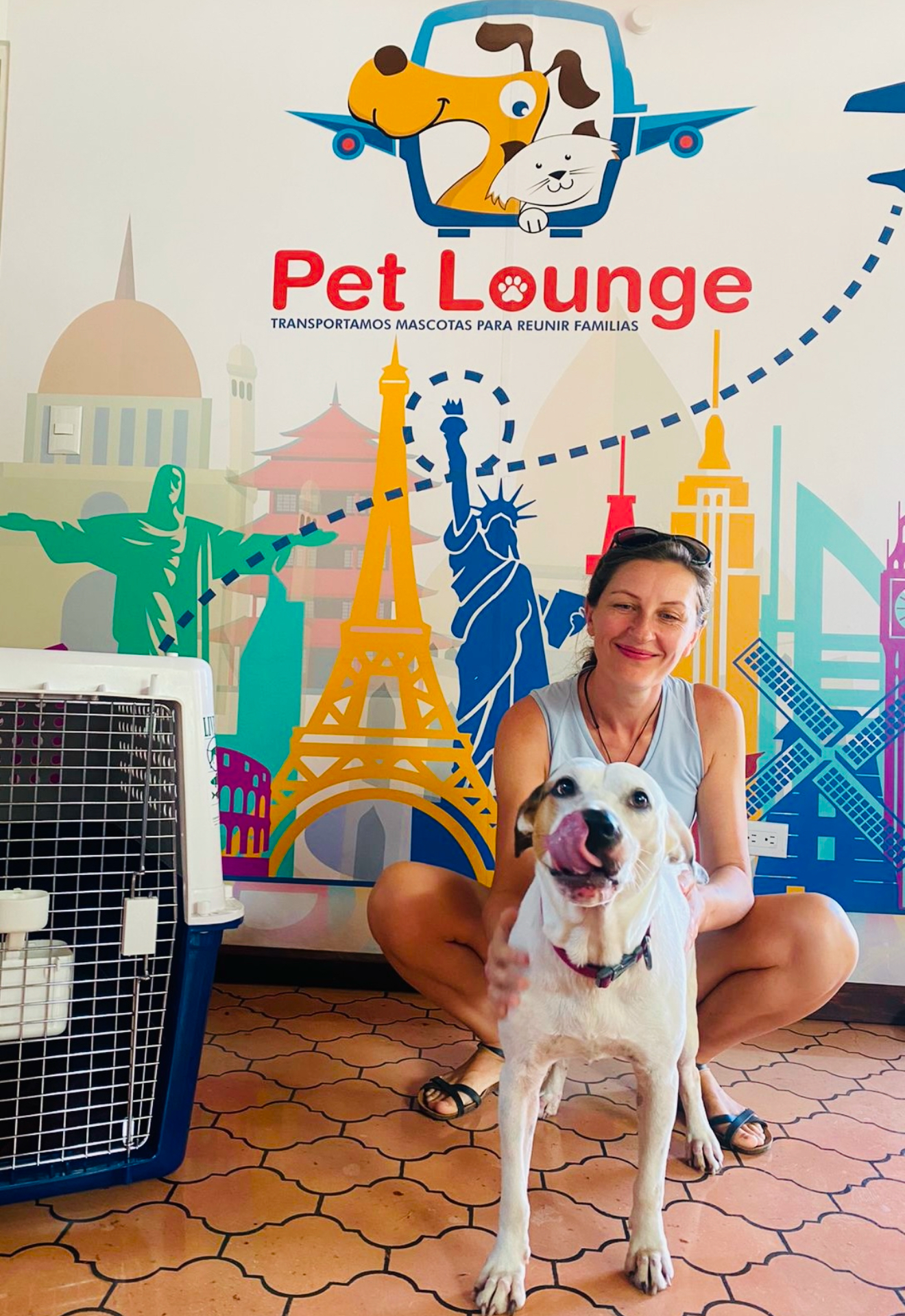 A woman posing with her dog in front of a colorful "Pet Lounge" mural, with a pet travel crate beside her.